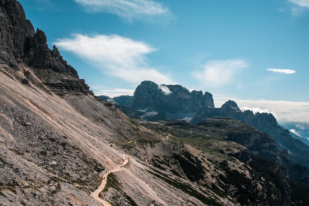 Blick-auf-den-Start-der-drei-Zinnenwanderung-oberhalb-der-Auronzohütte Blick auf den Start der drei Zinnen Wanderung oberhalb der Auronzohütte. Der Wanderweg schlängelt sich an dem Berg entlang. In der Ferne ist ein weitläufiges Gebirge zu sehen.