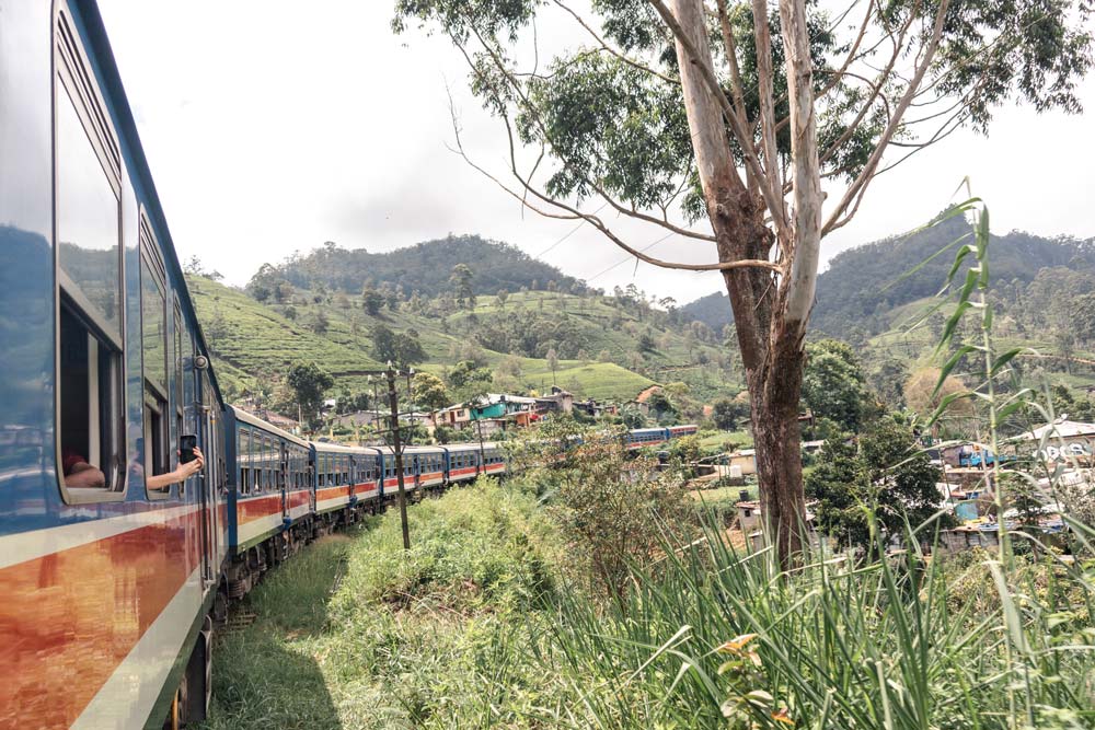 Auf-der-Zugstrecke-von-Kandy-nach-Ella-geht-es-durch-kleine-Dörfer-am-Fuße-der-Teehügel Zug auf der Strecke zwischen Kandy und Ella inmitten grüner Landschaft